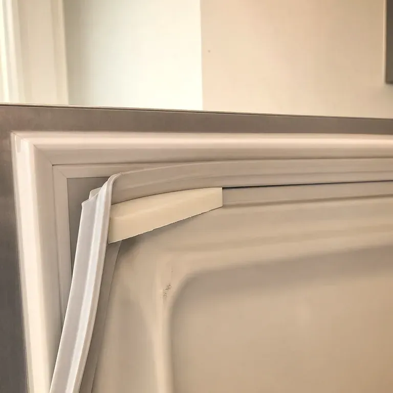 Top corner of a white refrigerator door gasket being eased out of its channel with a plastic wedge.