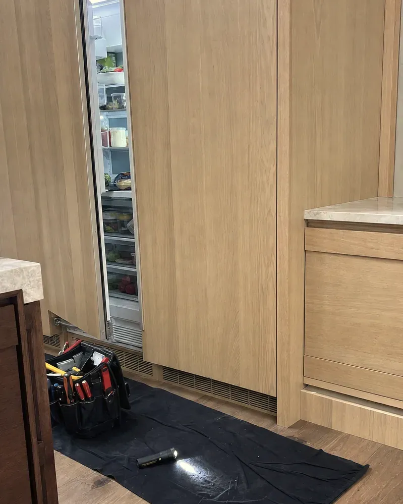 Panel-ready column in pale oak cabinetry standing open, a technician's tool bag and flashlight on a black mat in front.